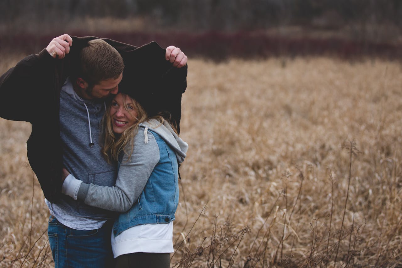 hero-img-02 A couple shares a warm embrace under a coat in a rustic autumn field, expressing love and togetherness.